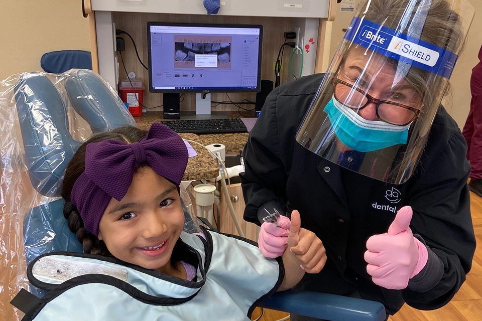 A dental hygienist and young girl giving thumbs up and smiles from dental chair.